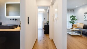A modern interior with a kitchen area featuring a sink and countertop, a hallway leading to a living room with a sofa and a coffee table, and a plant in the corner. - Bloom - Luxury 2-Bedroom Apartment in Athens (Greece - Glyfada)