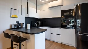 A modern kitchen with a black countertop, white cabinets, a stainless steel refrigerator, and a built-in oven. There are two black bar stools and a coffee machine on the counter. - Bloom - Luxury 2-Bedroom Apartment in Athens (Greece - Glyfada)