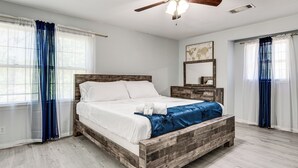 Une chambre à coucher avec un lit en bois, du linge de lit blanc, une table de chevet en bois, un miroir et un ventilateur de plafond.