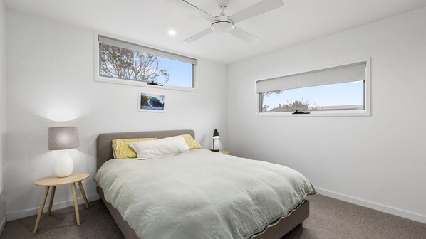 Un dormitorio con cama, dos mesitas de noche, ventilador de techo y dos ventanas con persianas.
