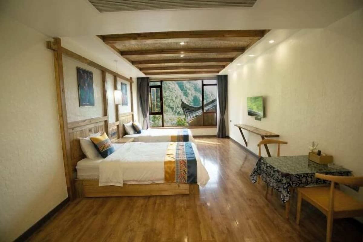 A room with two wooden beds, a dining table with chairs, a TV, and a view of a mountain.