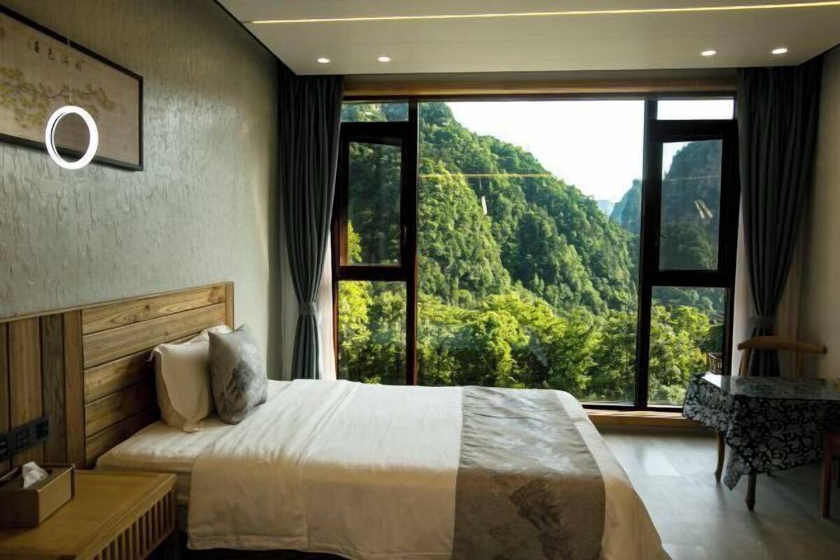 Un dormitorio con una cama grande, una mesita de noche, una pequeña mesa con una silla y vistas a una montaña boscosa.