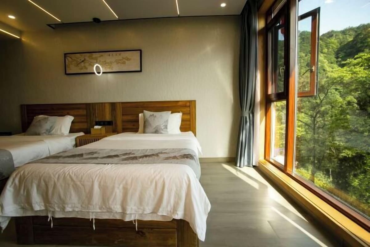 A bedroom with two beds, a wooden headboard, a large window with a forest view, and a wall-mounted clock.