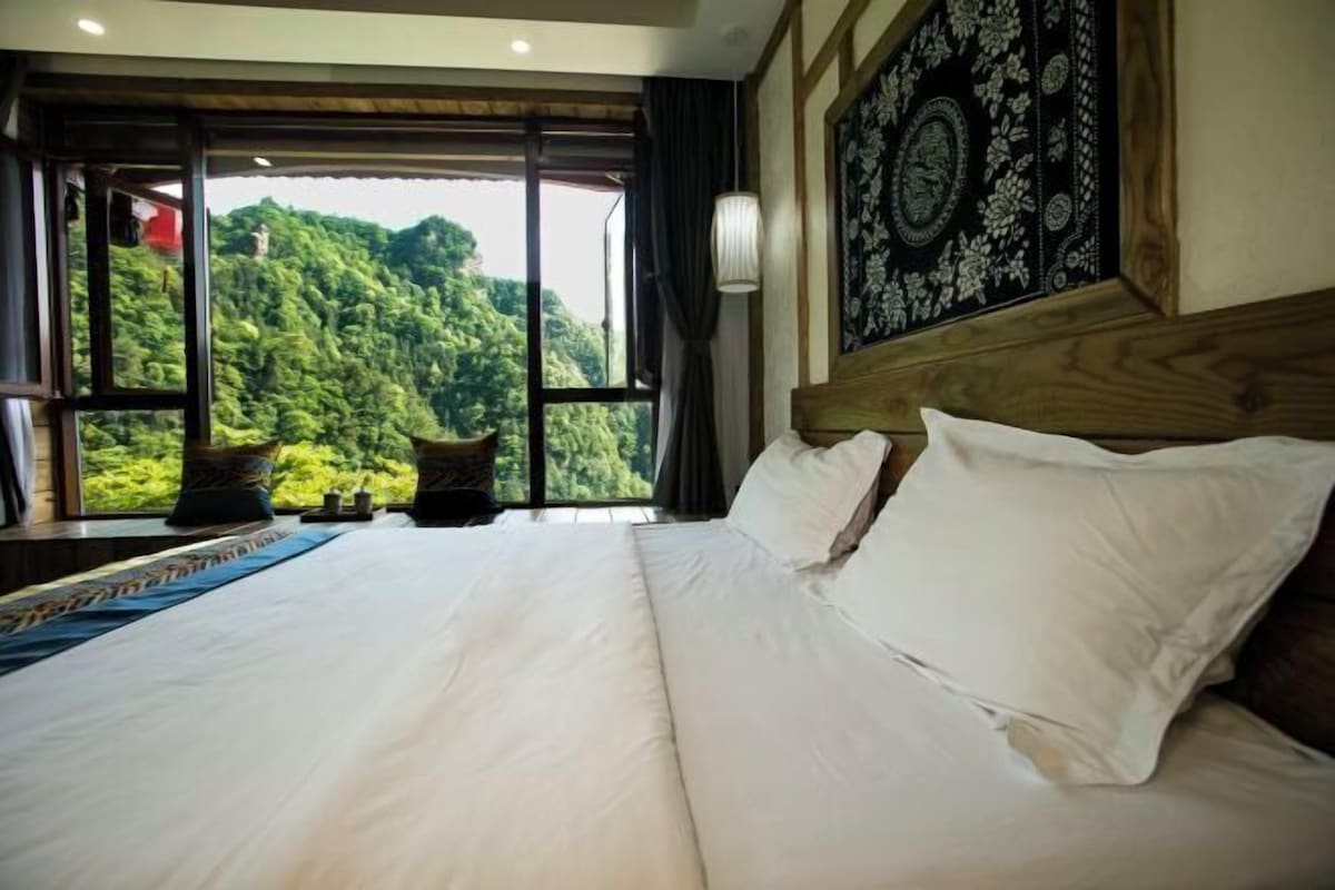 A bedroom with a large bed, a view of lush greenery, and a patterned tapestry on the wall.