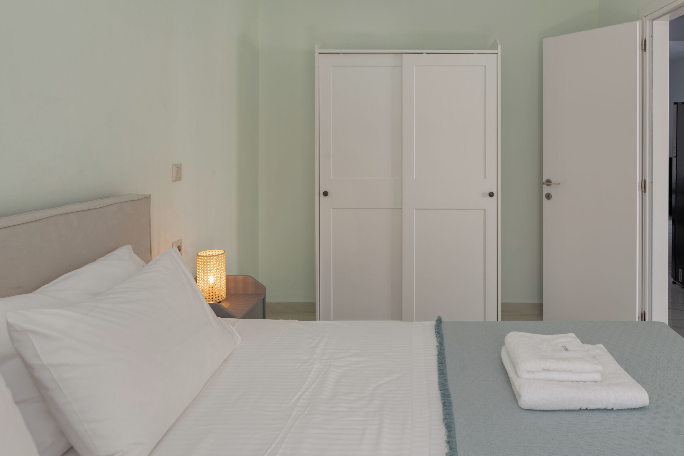 A neatly made bed with white linens, a bedside lamp, and a folded towel on a light-colored bedspread.