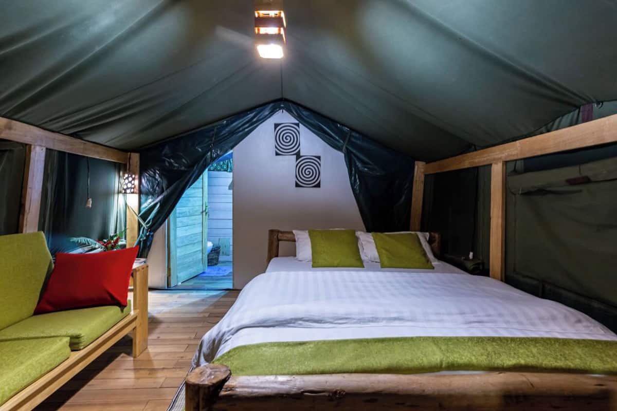 Una habitación tipo tienda de campaña, acogedora, con una cama, cojines verdes y un banco de madera.
