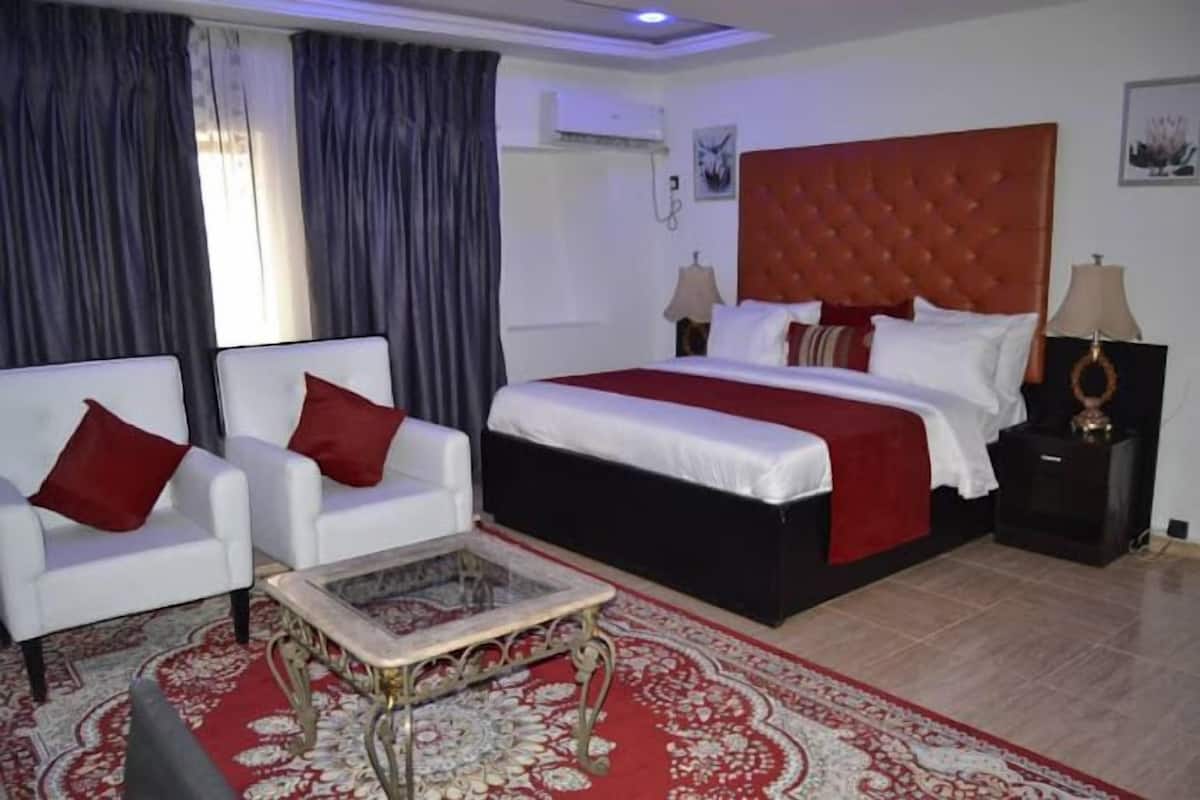 Habitación de hotel con una cama grande, dos sillones blancos, una mesita de centro pequeña y una alfombra roja estampada.