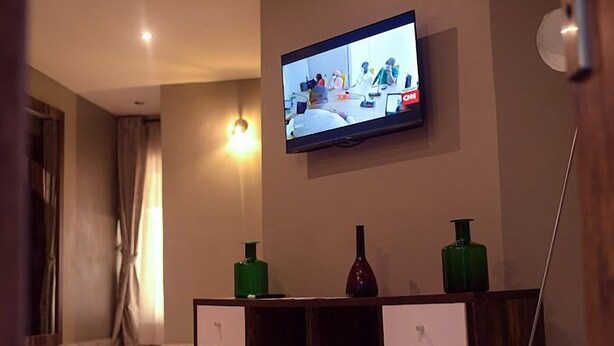 A modern living room with a flat-screen TV mounted on the wall, a wooden console table with drawers, and a vase. - La VOD Hotel (Benin City)