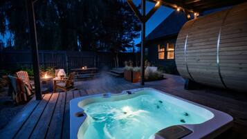 Un bain à remous installé sur une terrasse en bois, avec un espace salon couvert et un grand tonneau.