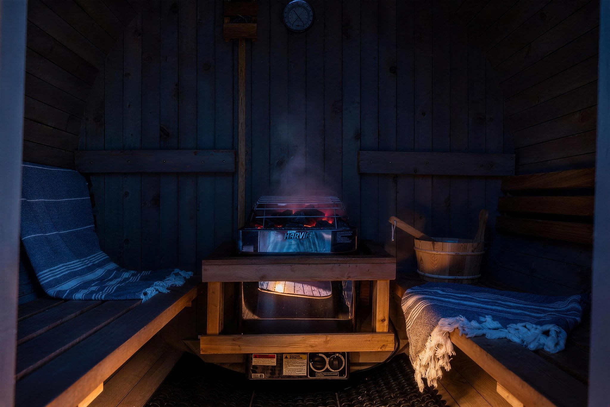 A sauna with a wooden bench, a bench with a blanket, and a heater emitting steam.