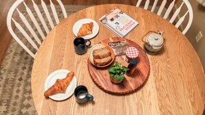 Een ronde houten tafel met een tijdschrift, een theepot, een kopje, een bord met een croissant, een bord met een stuk taart en een kleine bloempot met een plantje.