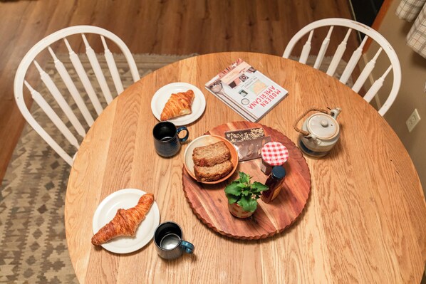 A round wooden table with a magazine, a teapot, a cup, a plate with a croissant, a plate with a slice of cake, and a small potted plant. - Cozy Airstream Sauna + Hot Tub | Sleeps 4 Downtown (Sandpoint)