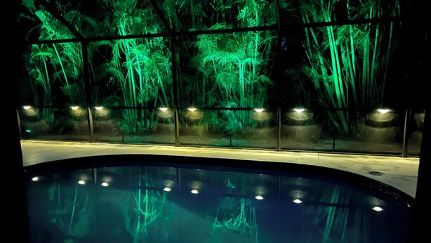 Ein Schwimmbad mit Glaswand, das das Wasser und eine hinter ihm liegende, grün beleuchtete Waldszene reflektiert.