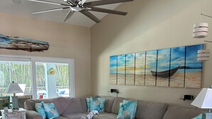 Ein Wohnzimmer mit Deckenventilator, einem großen Wandbild mit Strandmotiv, einer Sofagarnitur und einem Couchtisch.