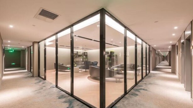 Un couloir d’hôtel moderne avec des cloisons en verre, un sol recouvert de moquette et un espace détente avec des canapés et une table basse.