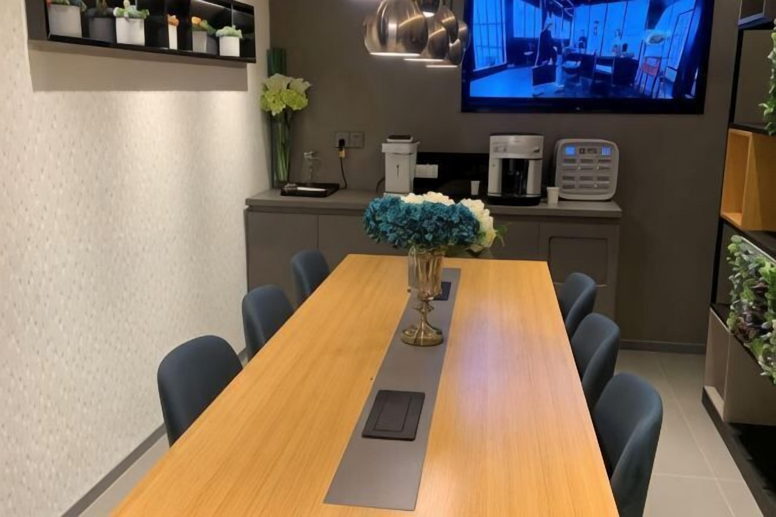 A conference room with a long table, chairs, a vase with flowers, a coffee machine, a TV, and shelves with plants.