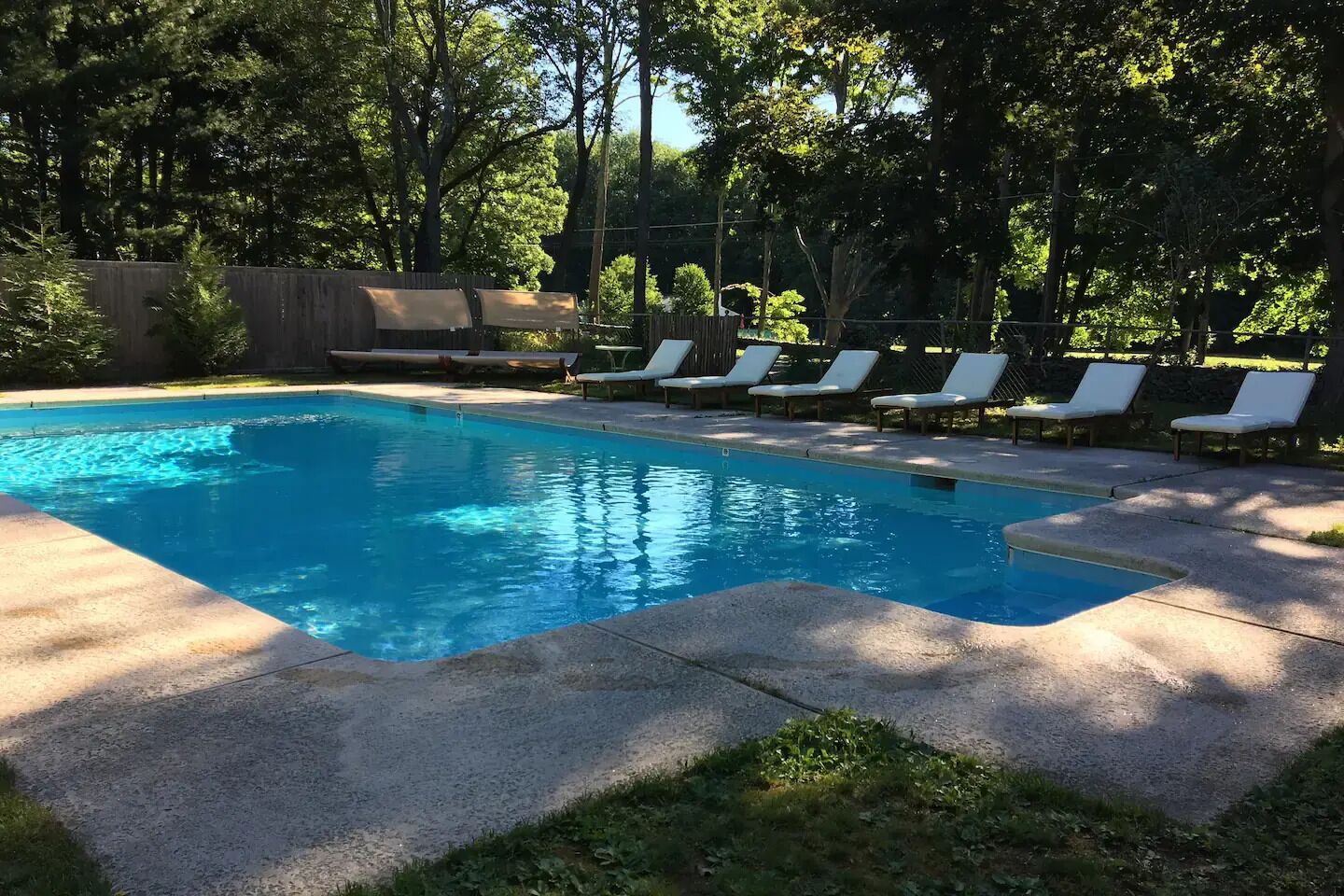 A rectangular swimming pool with a concrete edge, surrounded by a concrete deck. There are white lounge chairs aligned along the pool's edge.