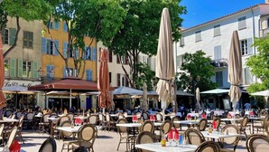 An outdoor café with tables and chairs, surrounded by buildings with shutters and awnings. - Casa-Jardin Secret: Apt climatisé avec piscine (Antibes-Juan-les-Pins)