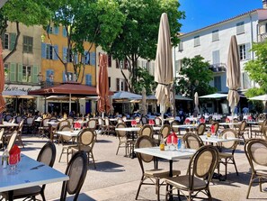 Un café al aire libre con mesas y sillas, rodeado de edificios con contraventanas y toldos.