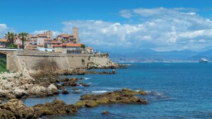 A coastal town with historic buildings, a stone wall, and a view of the sea and distant mountains. - Casa-Jardin Secret: Apt climatisé avec piscine (Antibes-Juan-les-Pins)
