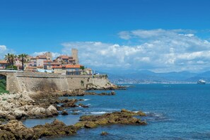 Un pueblo costero con edificios históricos, un muro de piedra y vistas al mar y montañas distantes.