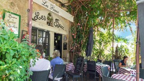 Un café al aire libre con mesas y sillas, un letrero que dice 'Les Baux' y una pizarra con el menú.