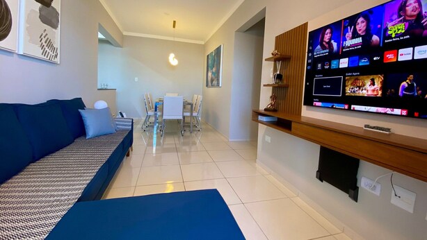 A living room with a sofa, a TV mounted on the wall displaying a streaming service, a dining area with a table and chairs, and a kitchen area with a countertop. - Edifício Breno Navarro  (Praia Grande)