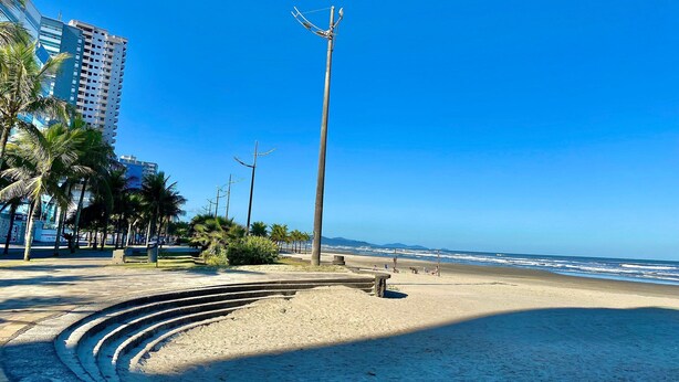 Una spiaggia con una struttura in cemento a forma di curva, palme e un cielo azzurro terso.