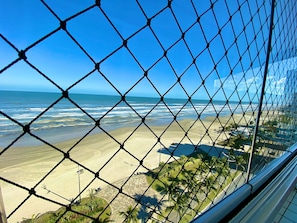 A beach view through a chain-link fence, including the ocean, sandy beach, and palm trees. - Edifício Breno Navarro  (Praia Grande)