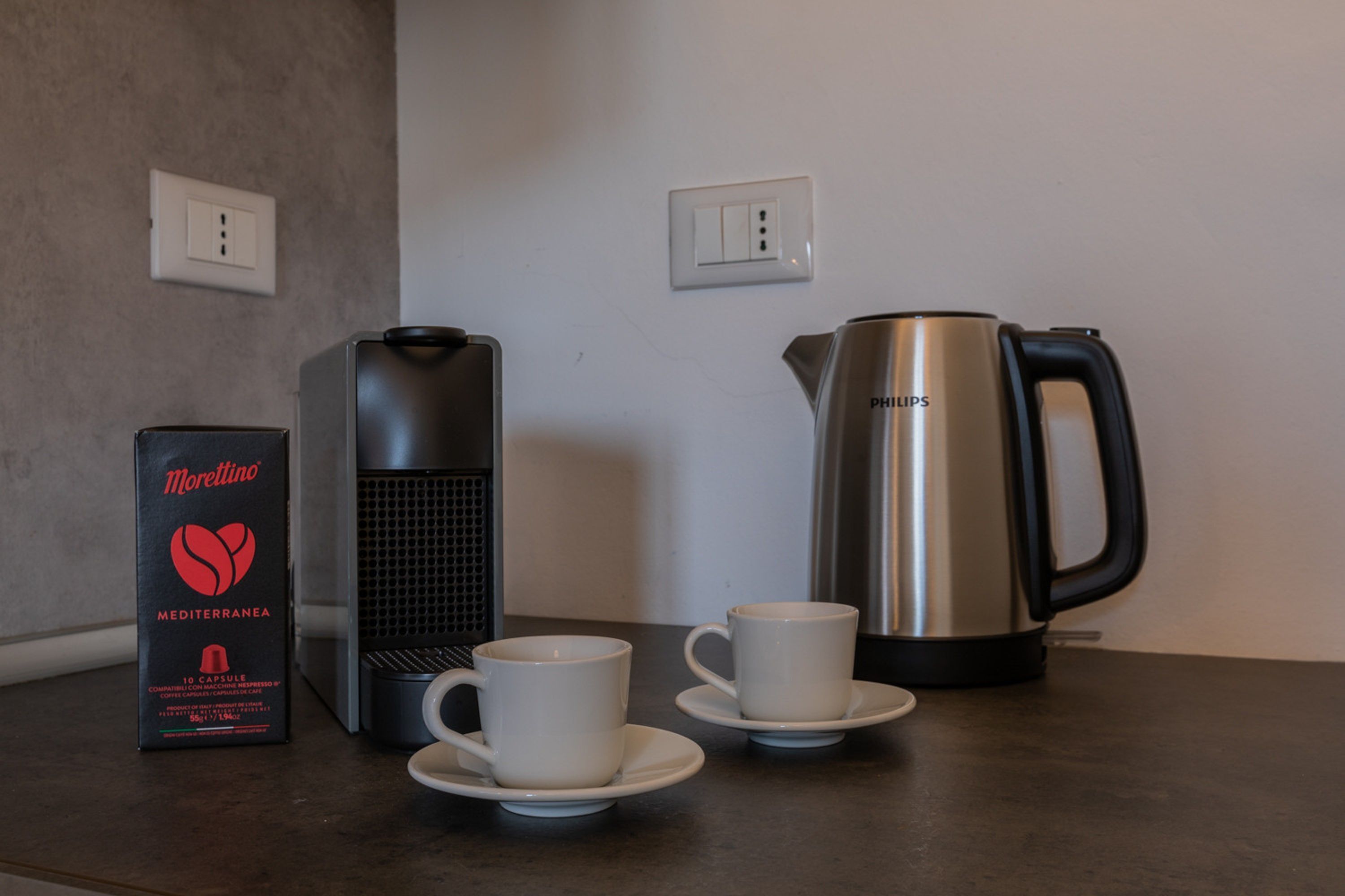 A coffee maker, a box of Morettino coffee capsules, and two white cups on a countertop.
