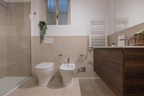 A modern bathroom with a glass shower enclosure, a toilet, a bidet, and a wooden vanity with a mirror.