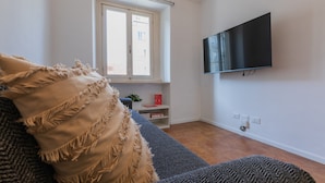 A living room with a sofa, a television mounted on the wall, a window with blinds, and a small shelf with books and a plant. - Il Papiro Apartment by Wonderful Italy (Turin)