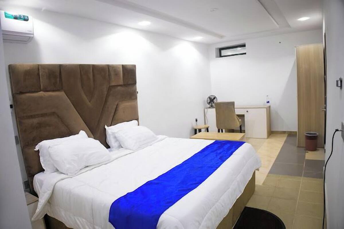 A neatly made bed with a blue accent, a wooden headboard, and a small desk with a chair in a modern room with white walls and a tiled floor.