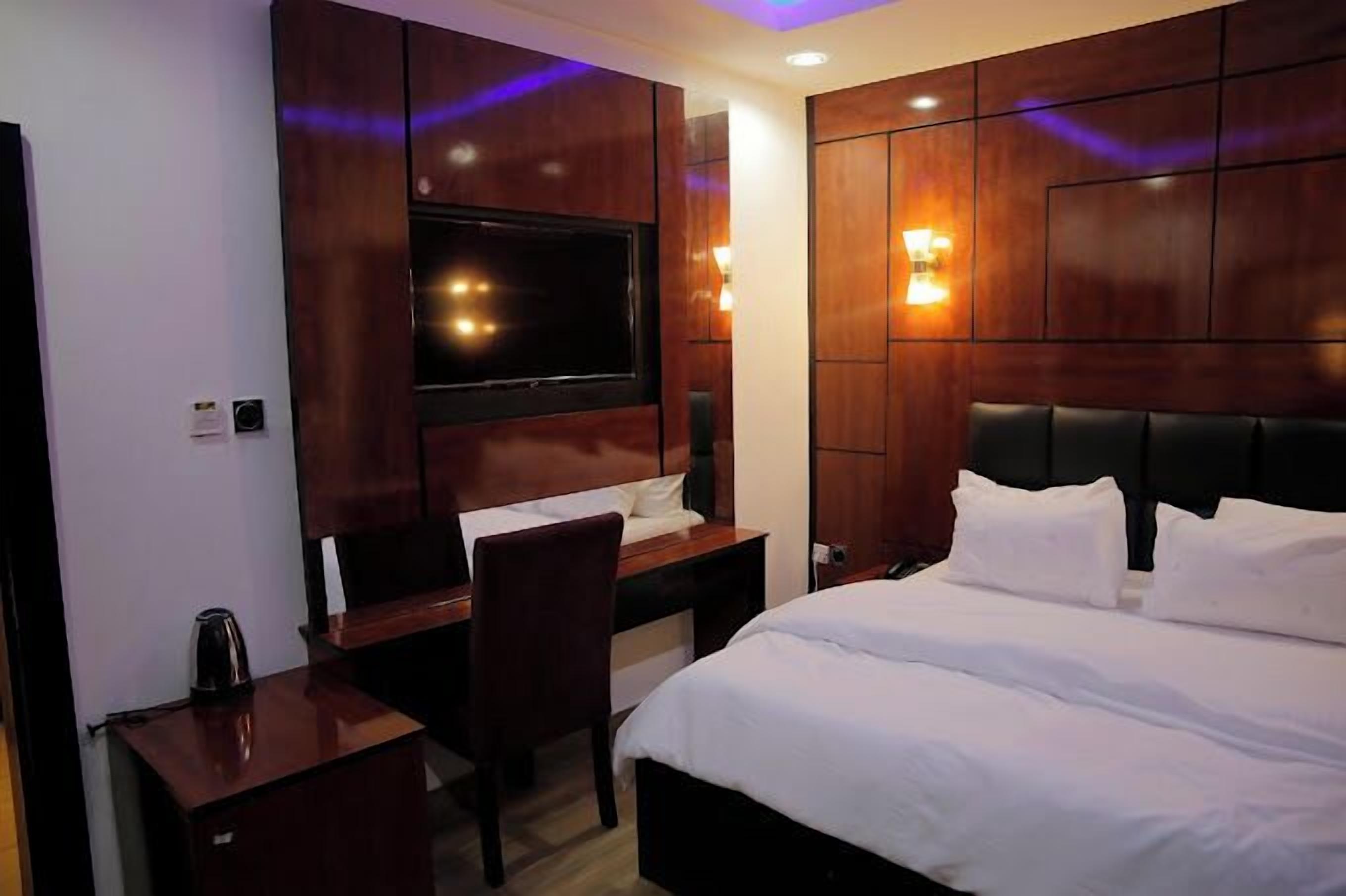 A hotel room with a bed, a desk, a chair, a TV, and wall-mounted lights.