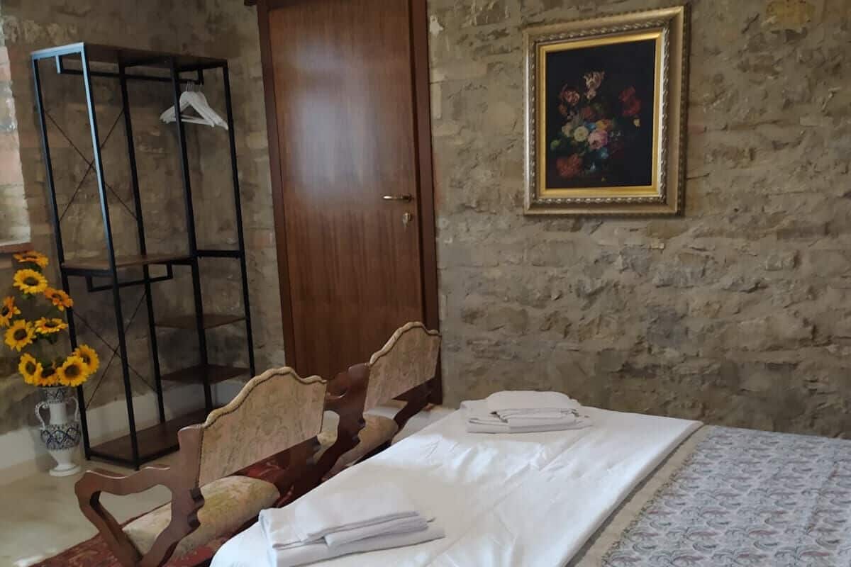 A bedroom with a bed, a wooden wardrobe, a chair, a small table with a vase of sunflowers, and a framed painting on the wall.