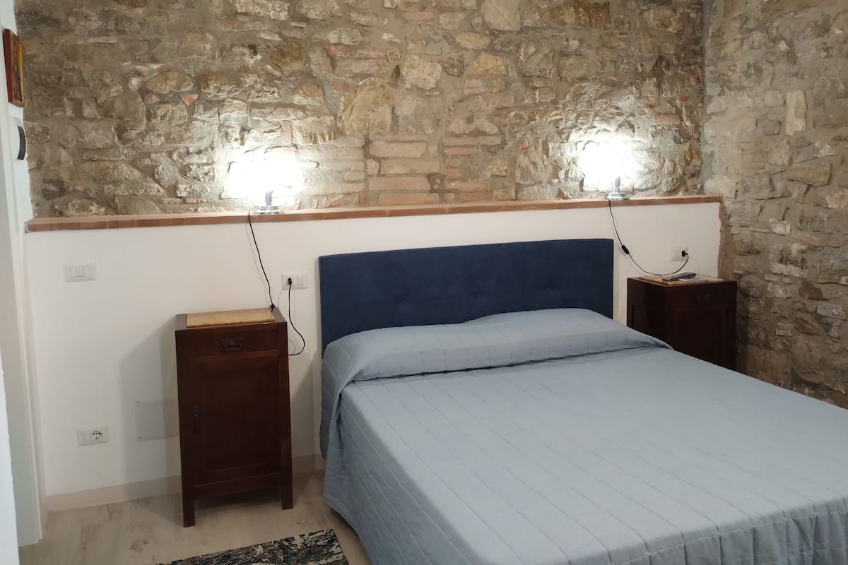 A bedroom with a stone wall, a blue headboard, a bed with a light blue bedspread, two bedside tables with lamps, and a small wooden nightstand.