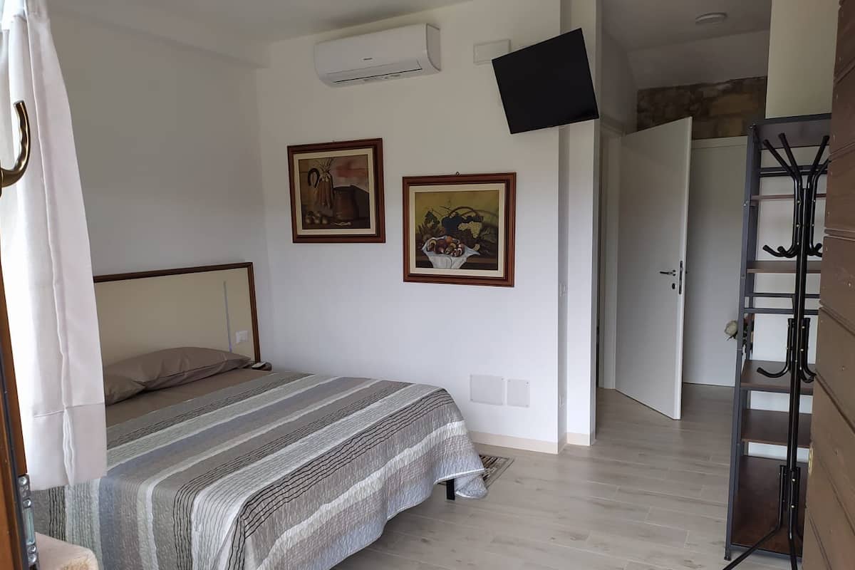 A bedroom with a bed, two framed pictures, an air conditioning unit, a TV mounted on the wall, and a wooden wardrobe with a coat rack.