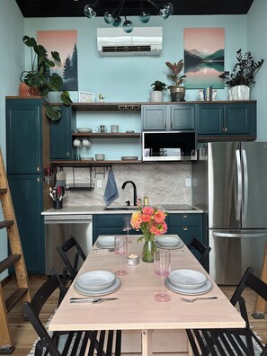 A modern kitchen with teal cabinetry, a stainless steel refrigerator, and a dining area set for two with a table, chairs, and a vase of flowers.