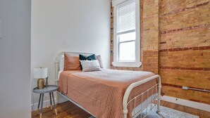 Une chambre à coucher avec un lit, une petite table de chevet, une lampe, une fenêtre et un mur en briques.