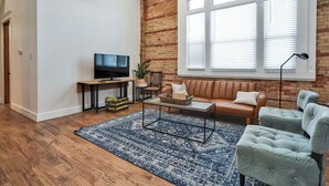 Un salon avec un mur en briques, une grande fenêtre avec des stores, un ventilateur de plafond, un téléviseur, un canapé, une table basse et un tapis.