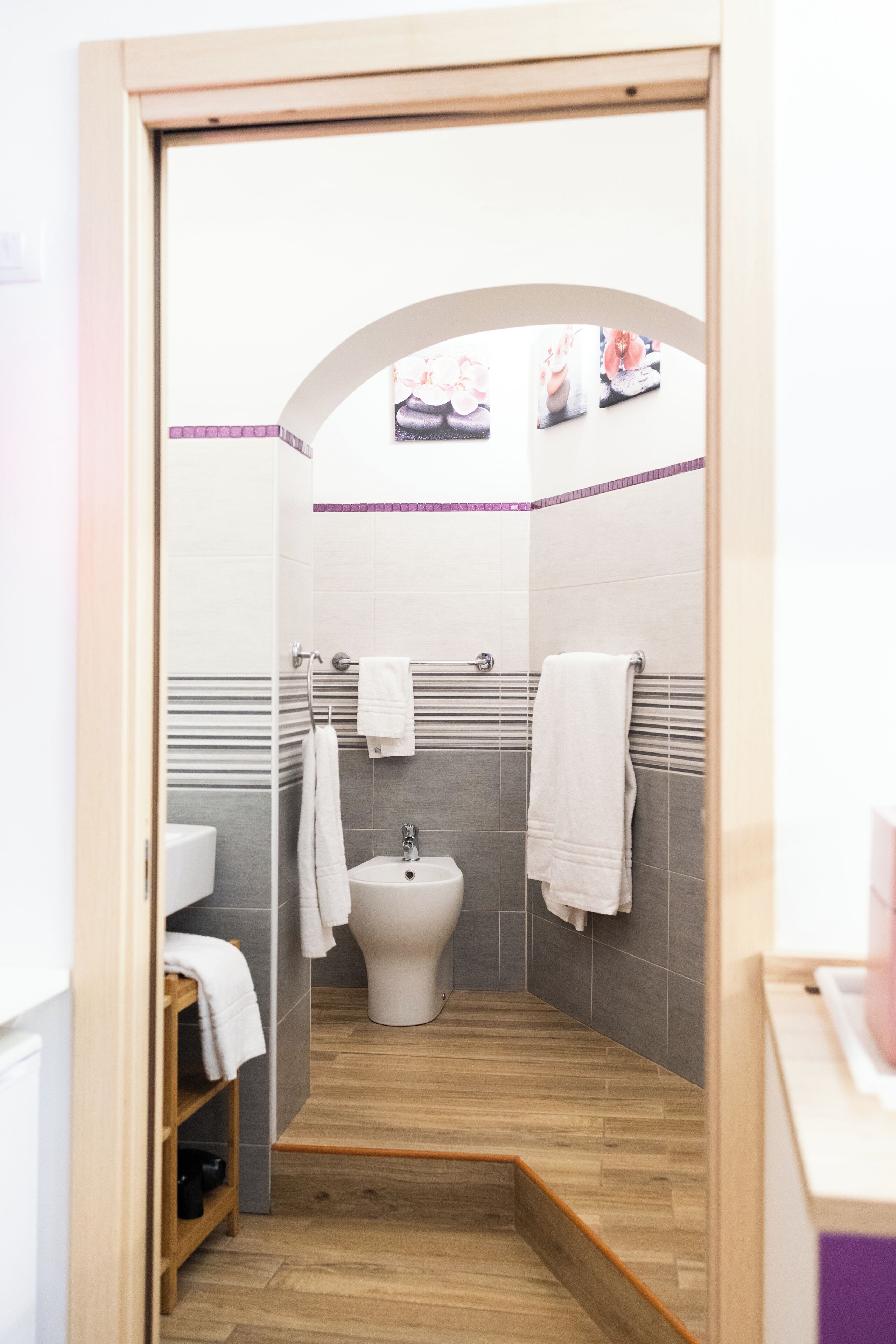 A small, modern bathroom with a shower, toilet, and sink. There is a wooden shelf with towels and a small window above the sink.