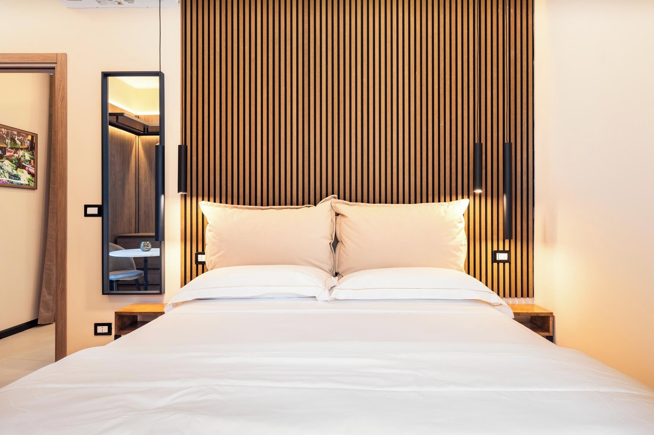 Una cama bien hecha en una habitación de hotel con cabecera de madera, una mesita con una silla y un espejo en la pared.