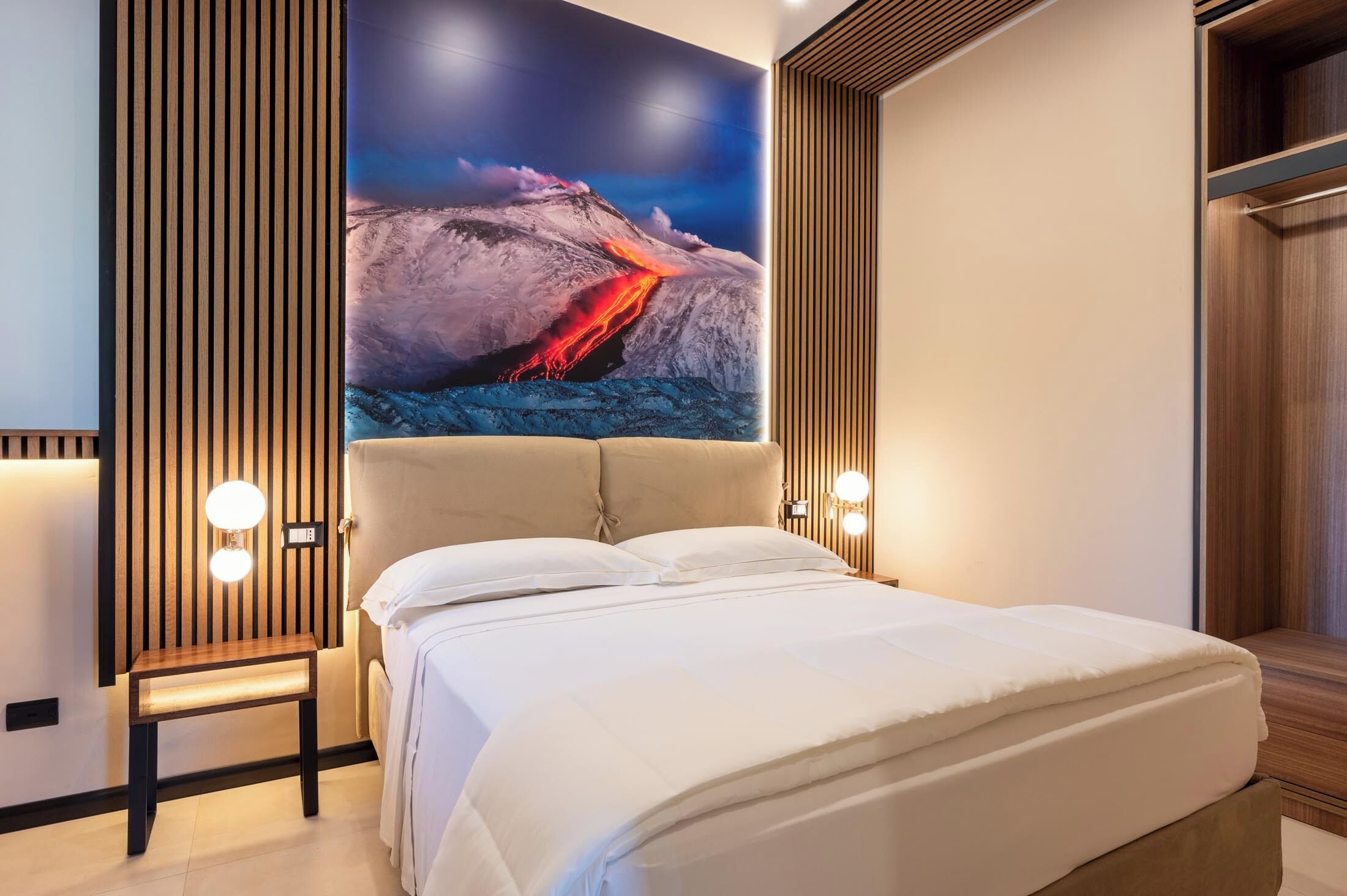 A modern hotel room with a large bed, a wooden headboard, and a wall-mounted artwork depicting a volcanic eruption.