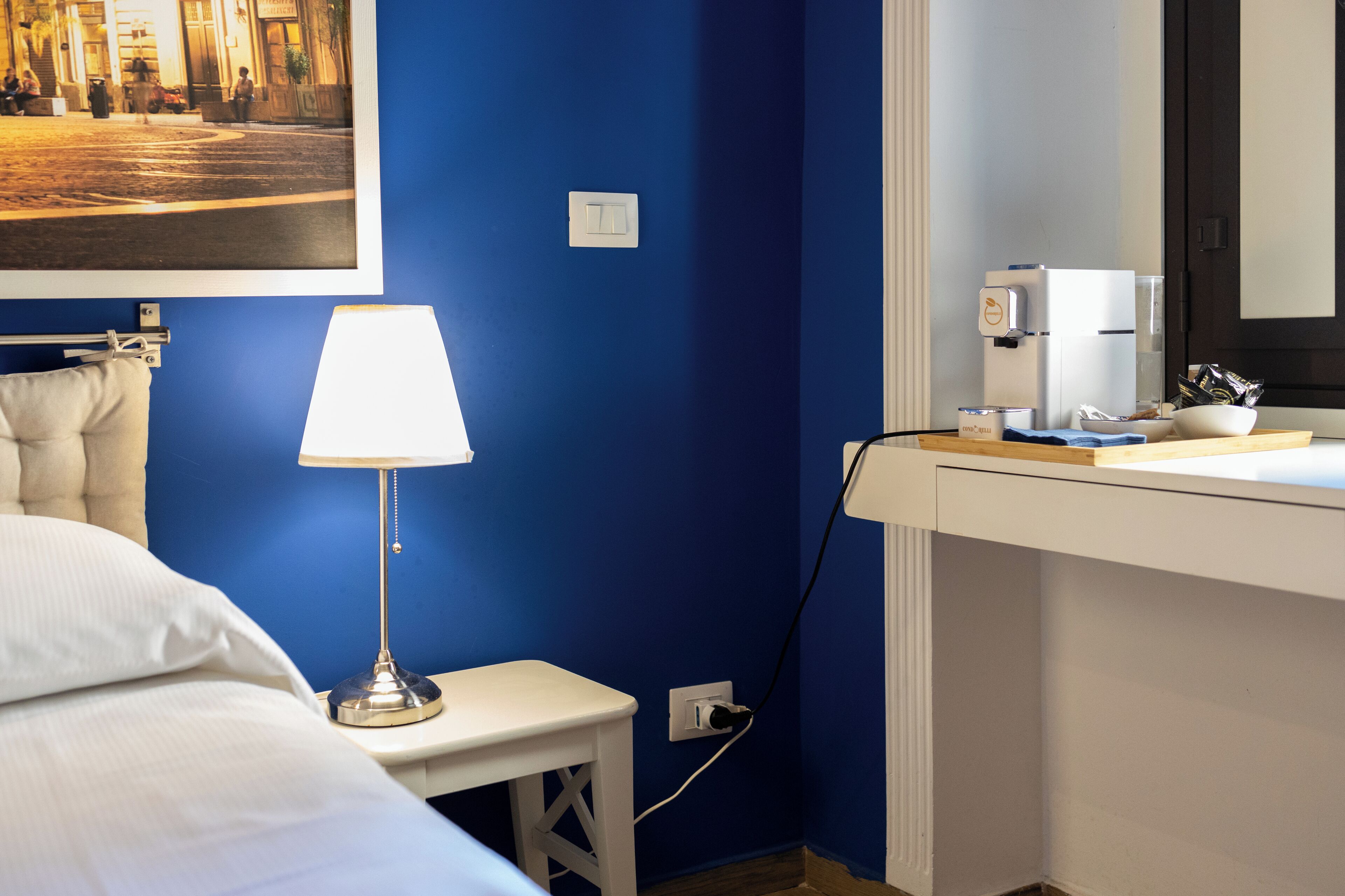 Ein Hotelzimmer mit einem Bett, einem Nachttisch mit Lampe, einem kleinen Hocker, einem weißen Schreibtisch mit Kaffeemaschine und einem gerahmten Bild an der Wand.