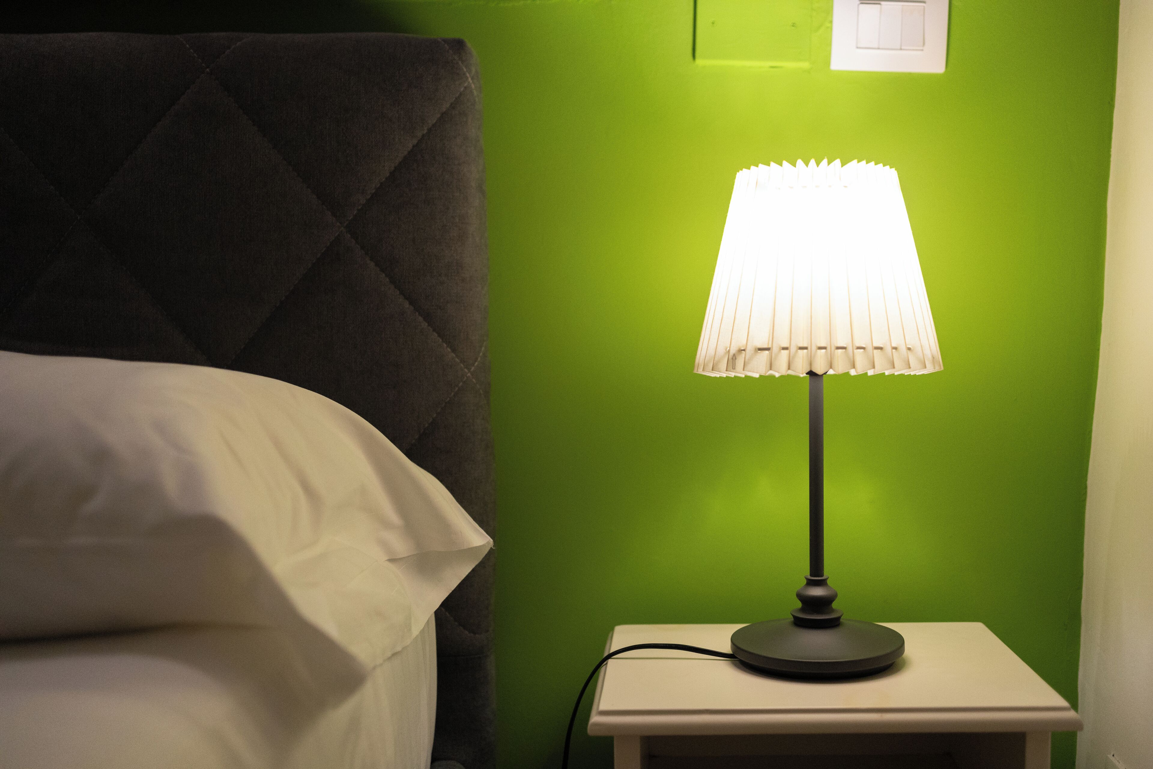 A bed with white linens, a dark headboard, and a bedside table with a lamp against a green wall.