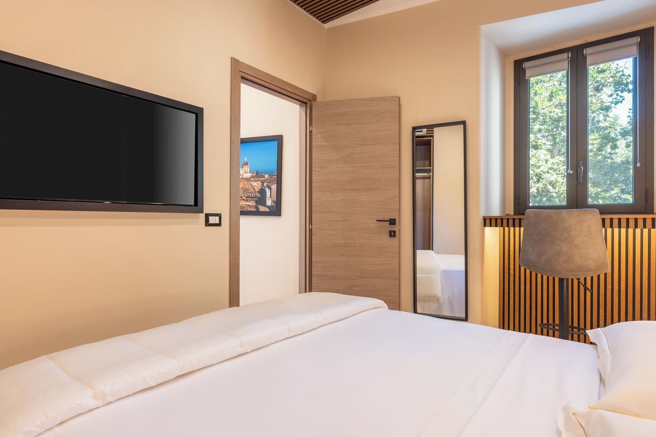 A modern hotel room with a large bed, a flat-screen TV mounted on the wall, a wooden door, and a window with blinds.