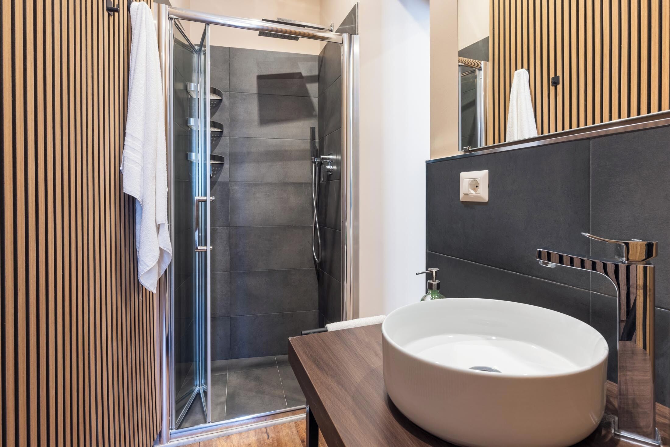 A modern bathroom with a glass shower enclosure, a wooden vanity with a white sink, and a wooden panel wall.