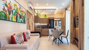 Un salon moderne avec un canapé, des coussins colorés, un coin repas avec des chaises, une cuisine avec des placards et un grand tableau représentant un cactus accroché au mur.