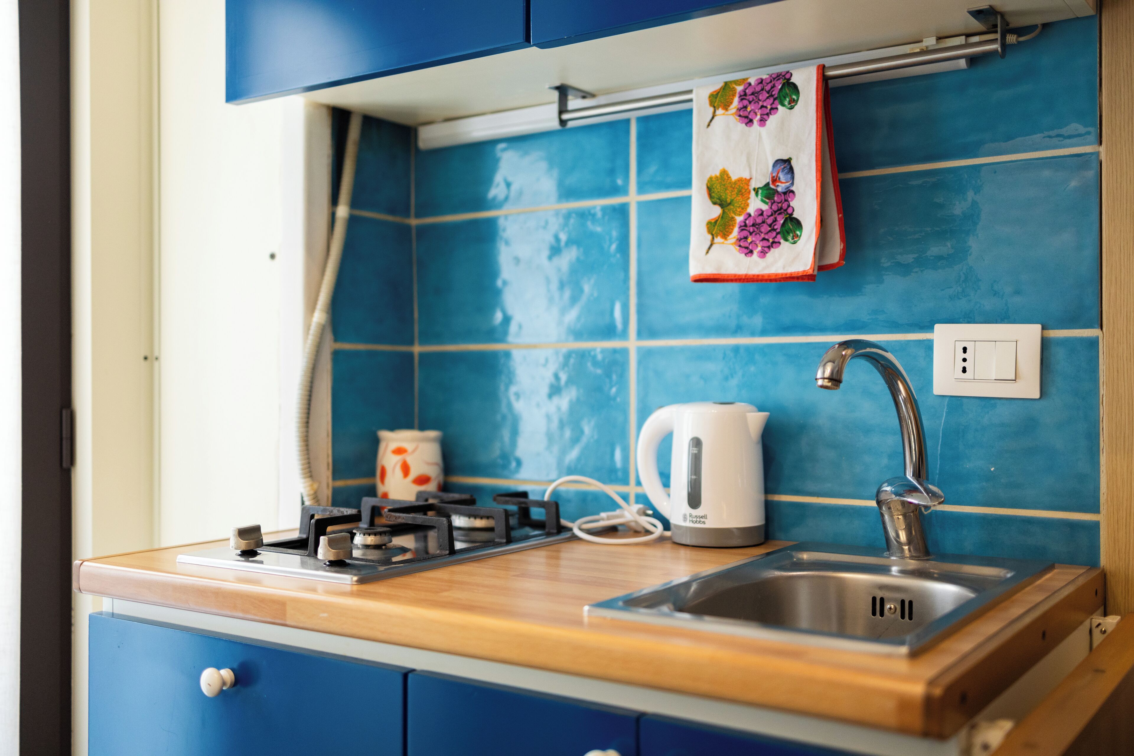 A kitchen with blue tiles, a wooden countertop, a kettle, a stove, and a sink.
