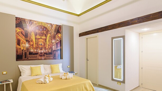 A hotel room with a large bed, a bedside table, a nightstand, a wall-mounted mirror, and a door. - Barlaman Luxury Rooms (Palermo)
